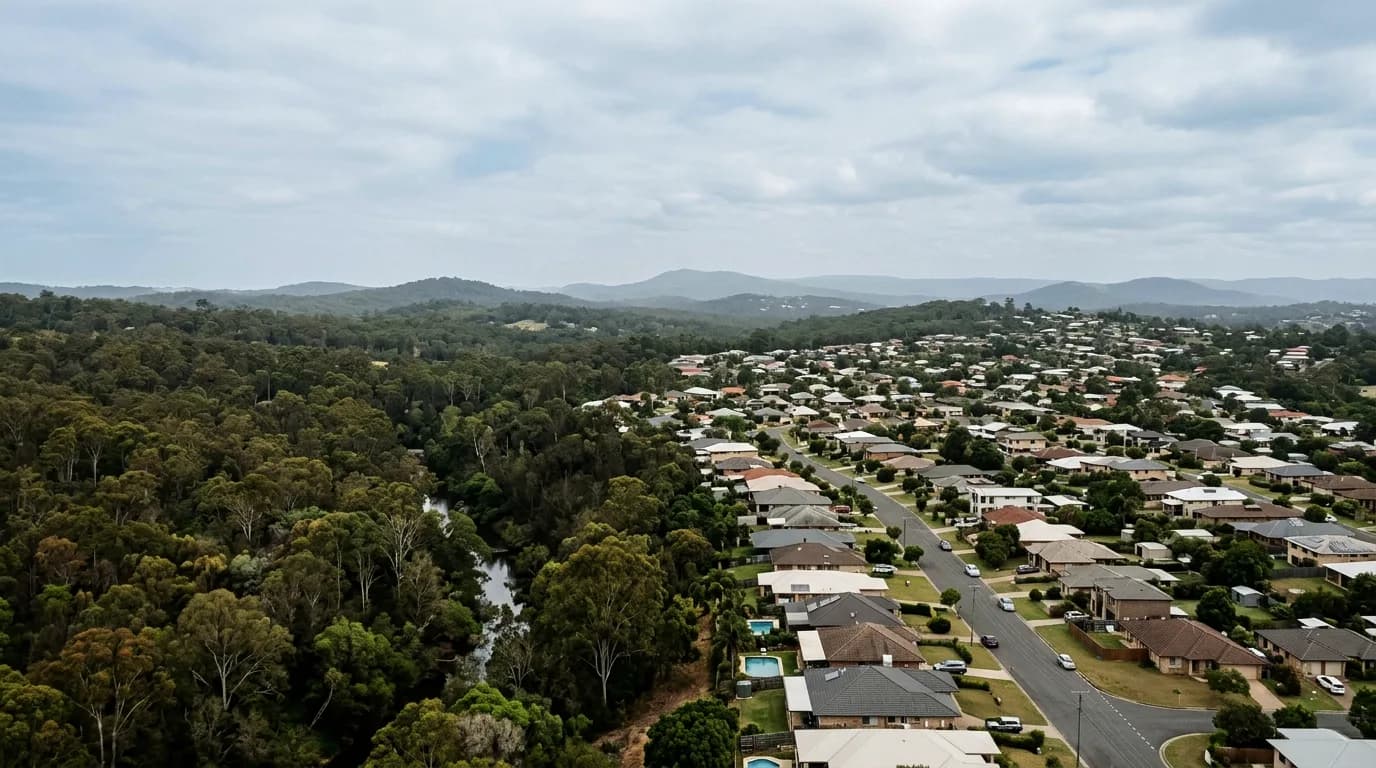 An elevated view across South East Queensland where established suburban rooftops meet dense remnant bushland along a creek line, low hills receding into haze beyond